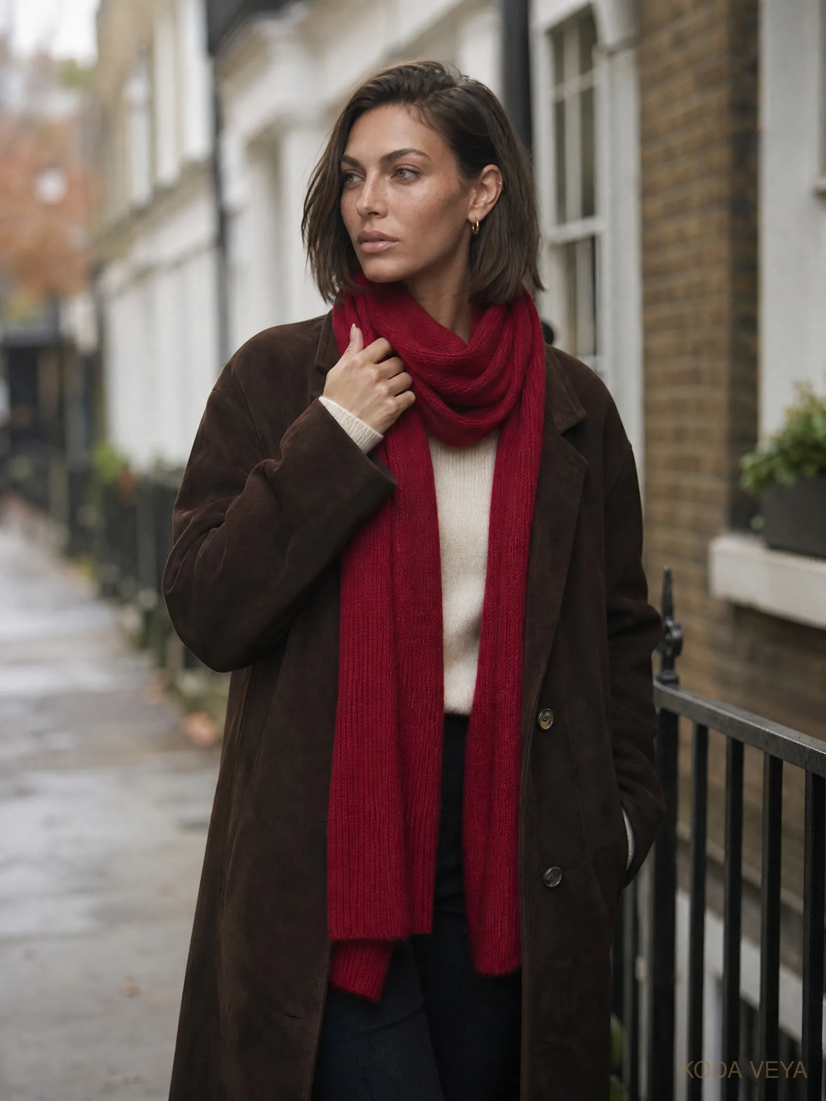 Koda on a London street in a long chocolate coat and cream top, wrapped in a cherry-red knit scarf
