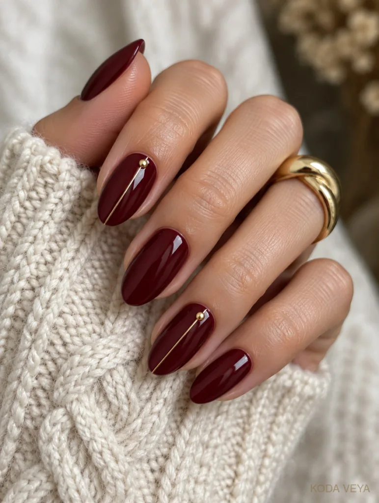 Glossy cherry-burgundy nails with a fine gold line on two nails, against a cream cable knit